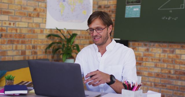 Male teacher smiling while using laptop in a modern classroom with exposed brick walls. Ideal for illustrating online teaching, e-learning, and modern education technology. Useful for educational websites, teacher training programs, and online course promotions.