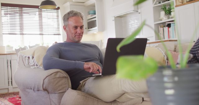 Happy caucasian man sitting on sofa in living room, using laptop - Download Free Stock Photos Pikwizard.com