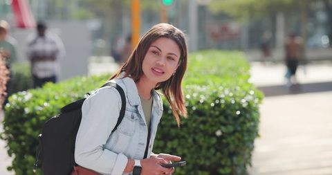 Young woman carrying backpack checking smartphone standing by green hedge on city sidewalk