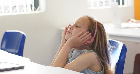 Schoolgirl daydreaming while resting chin in hands at sunlit classroom desk near laptop
