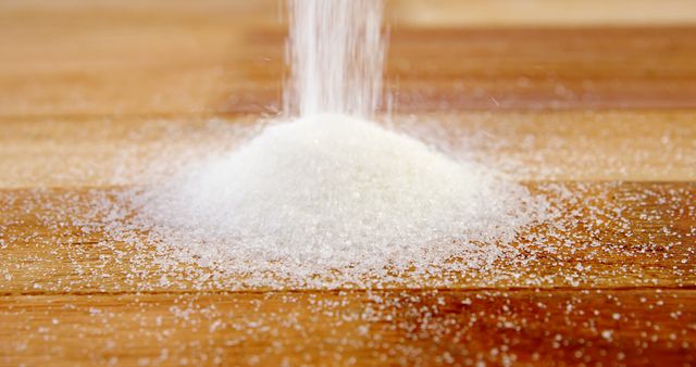 Sugar is being poured onto a wooden surface, creating a small pile with granules scattered around. Highlighting the texture and simplicity, the image captures a common ingredient used in cooking and baking.