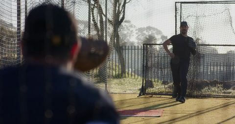 Mature men practicing pitching and catching in netted batting cage with l-screen, sunlight