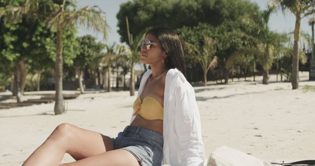 Young woman relaxing on a sunny beach wearing a yellow bikini top, denim shorts, white covering, and sunglasses. Palm trees and sand give a tropical vibe. Ideal for use in vacation travel promotions, summer lifestyle advertisements, and beachwear fashion marketing.