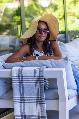 Woman relaxing on sunlit patio sofa wearing wide-brim straw hat and stylish sunglasses
