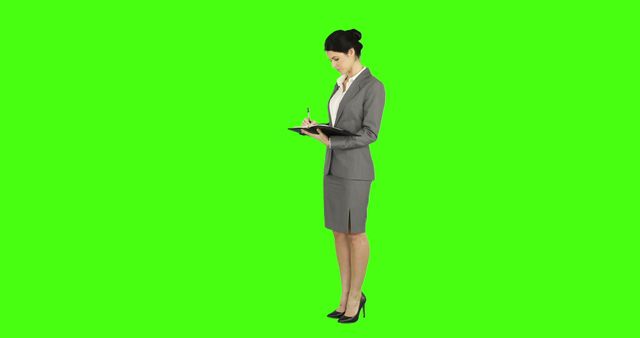 A professional Asian businesswoman is focused on writing notes on a clipboard, with copy space. Her attire suggests a formal business environment, and the green screen background allows for various graphical customizations.