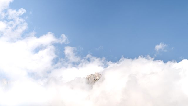 Mountain peak partially covered by thick, fluffy clouds against a clear blue sky. Ideal for backgrounds, travel promotions, outdoor adventure blogs, and inspirational nature content.