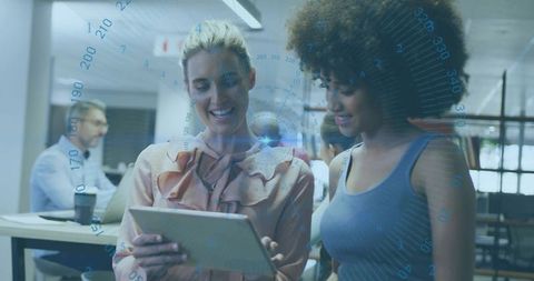 Female colleagues collaborating over tablet with futuristic hud overlay in modern office