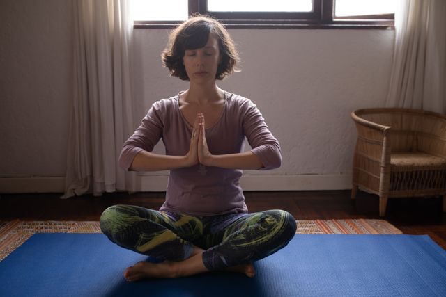Front view of a Caucasian woman with short dark hair, wearing sports clothes, sitting on the yoga mat with eyes closed in yoga position.