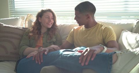 Relaxed interracial couple sitting on sunlit sofa holding hands sharing warm conversation