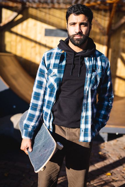 Biracial man holding skateboard in skate shop, wearing plaid shirt and hoodie. Ideal for use in advertisements for skateboarding gear, urban lifestyle promotions, and youth culture campaigns.