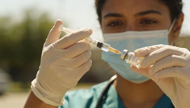 Nurse wearing surgical mask drawing vaccine from vial with syringe outdoors in sunlight
