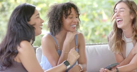 Diverse women laughing together on outdoor sofa sharing smartphone and smartwatch fun