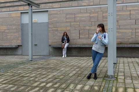 Young woman leaning on column drinking coffee in urban plaza while friend checking phone