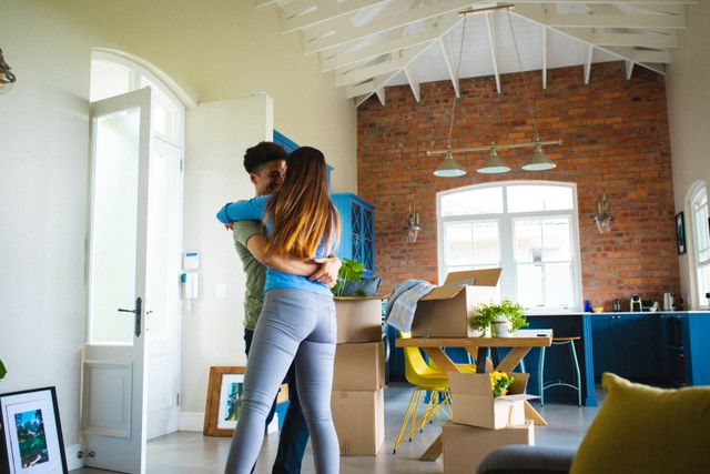 Young multiracial couple embracing and dancing in their living room, surrounded by moving boxes and modern decor. Ideal for use in articles or advertisements about moving into a new home, relationships, lifestyle, or home decor.