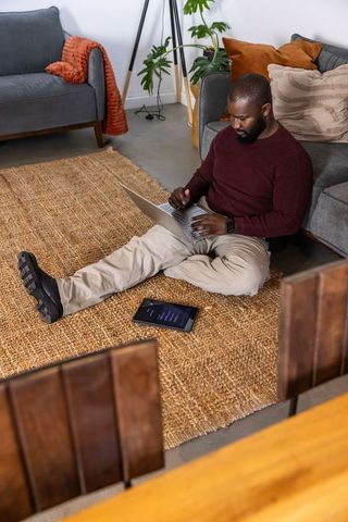African american man working from home on laptop sitting on jute rug in cozy living room