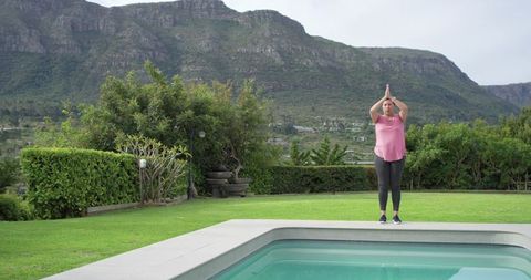 Mature woman practicing yoga poolside on manicured lawn with mountain backyard vista