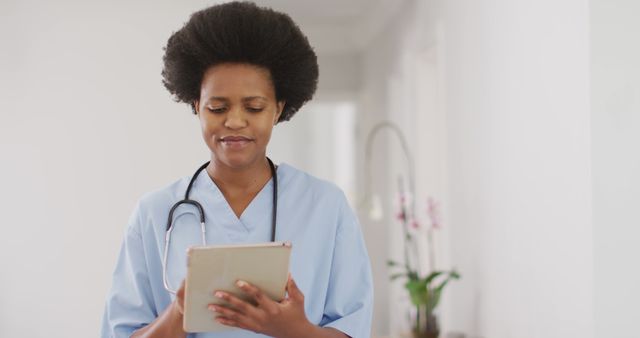 African American female nurse wearing scrubs and stethoscope, using a digital tablet for patient data entry. Ideal for healthcare industry websites, nursing programs, hospital promotional material, or technology in healthcare articles.