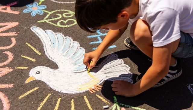 Boy kneeling drawing white chalk dove with sun rays on asphalt during sunny outdoor play