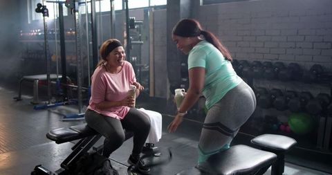 Diverse women laughing and hydrating during gym break in weight room friendly workout