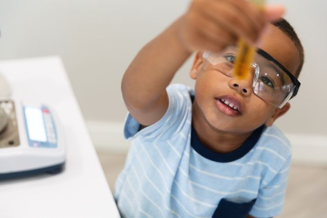 African american elementary boy wearing protective eyewear while looking at chemical in test tube. unaltered, education, laboratory, stem, scientific experiment, protection and school concept.