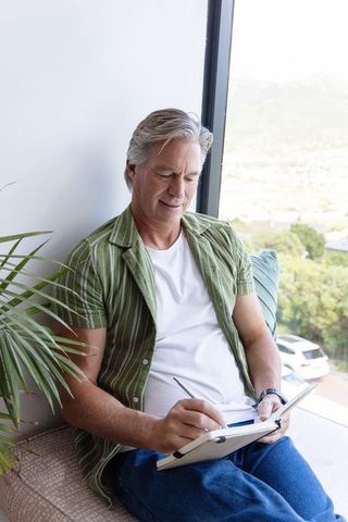 Mature man journaling on sunlit window seat overlooking hillside view