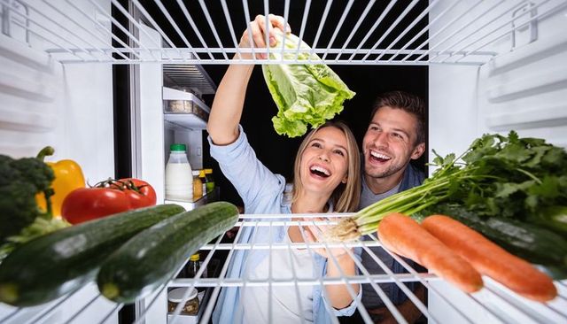 Happy couple arranging fresh vegetables in open refrigerator for healthy meal prep