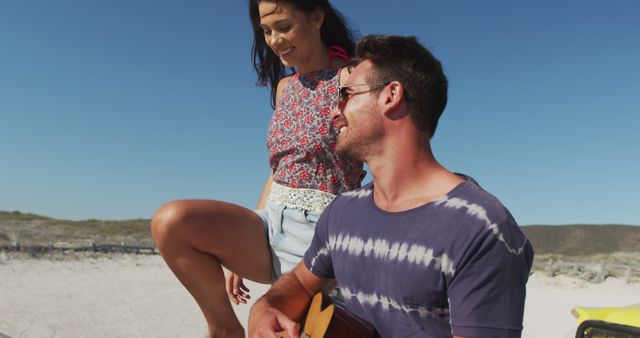 Couple relaxing on a sunny beach, guy playing guitar while the girl smiles and enjoys the moment. They are casually dressed, with the beach and ocean providing a scenic backdrop. Ideal for themes of summer vacations, leisure activities, youthful romance, happiness, and outdoor enjoyment. Perfect for travel blogs, lifestyle articles, and vacation advertisements.