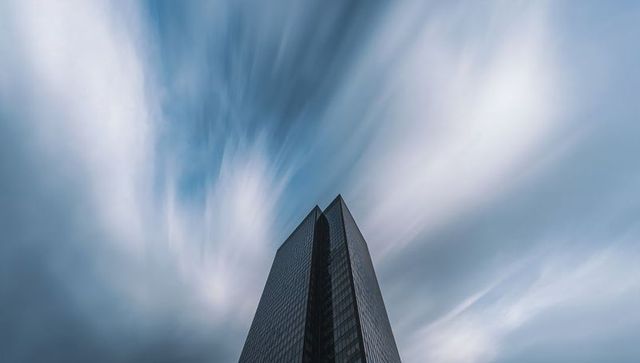 Rising glass skyscraper piercing dramatic sky with streaking clouds and reflective facade
