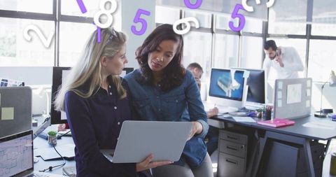 Female colleagues collaborating on laptop with animated numeric overlay in open office