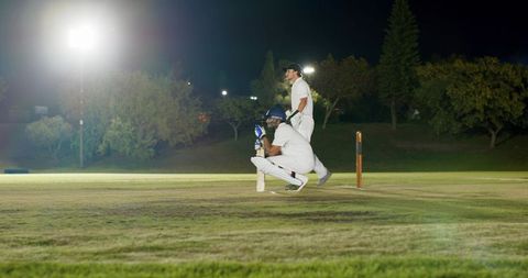 Night cricket teammates crouching and standing at crease under floodlights holding bat by stumps