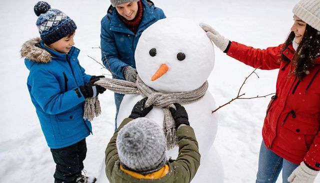 Family building snowman tying knit scarf placing stick arms smiling kids in winter jackets