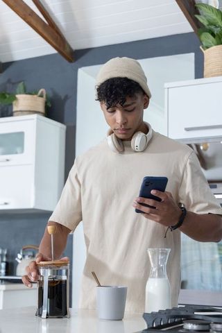 African american man making french press coffee while checking phone in modern kitchen