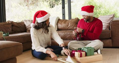 Diverse couple wrapping christmas gifts wearing santa hats in cozy sunlit living room