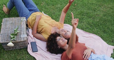 Two women lying on picnic blanket pointing at sky with wicker basket and smartphone