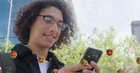 Young Man Engaging with Smartphone Amid Digital Emojis Outdoors