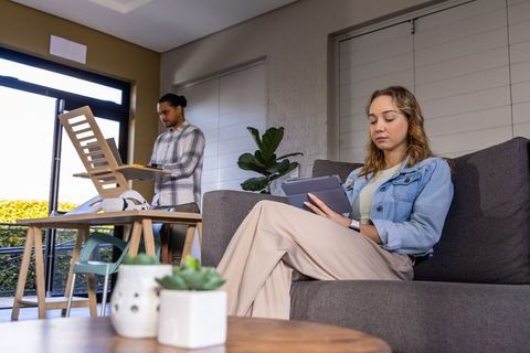 Couple multitasking: woman reading tablet on sofa while man assembling chair in sunlit living room