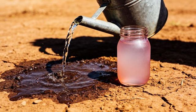 Pouring water from metal watering can into muddy puddle on cracked red soil with pink mason jar