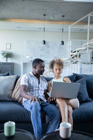 Diverse couple sitting on couch sharing laptop together in modern minimalist living room