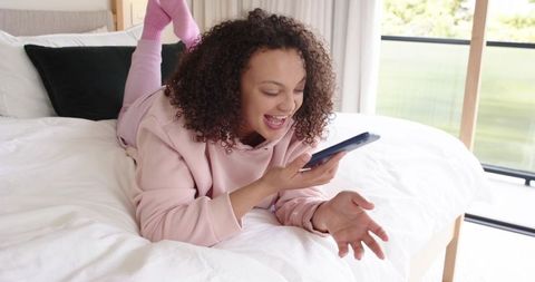 African american woman laughing while videochatting on smartphone lying on cozy white bed