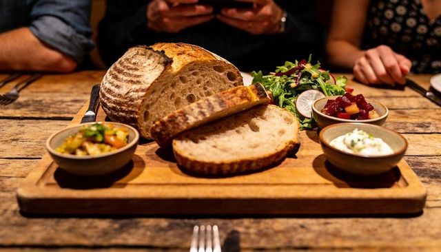 Slicing artisan sourdough loaf on rustic wooden board with condiments and greens