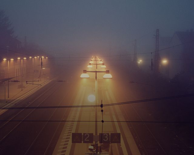 The image captures an empty train station platform shrouded in thick fog and darkness with glowing lamps illuminating the scene. Ideal for use in materials related to travel, transportation, night photography, weather conditions, or urban environments. This image conveys a sense of solitude, mystery, and calm.