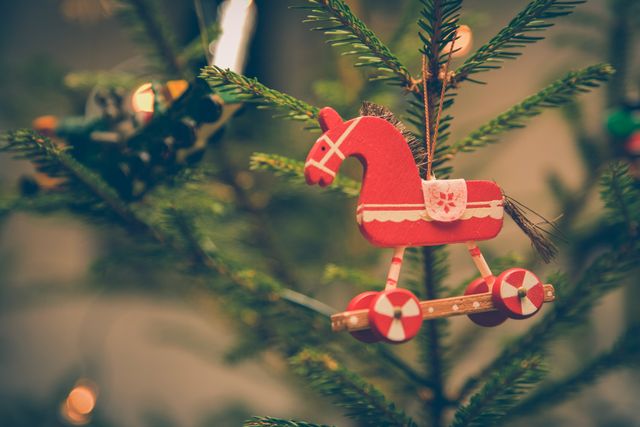 Vintage red wooden horse ornament hanging on Christmas tree branch with holiday lights glowing in background. Perfect for Christmas cards, festive social media posts, holiday decorations inspiration.
