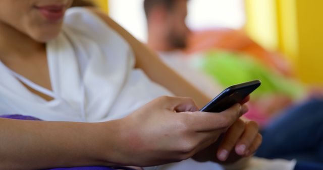 This image presents a young woman relaxing at home while using her smartphone. A blurred figure is seen in the background. Useful for themes related to technology use, relaxation, and lifestyle. Appropriate for articles, blog posts, and marketing materials focused on social media, casual activities, or the modern use of mobile devices.