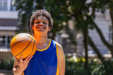 African american man holding basketball smiling on sunlit outdoor court