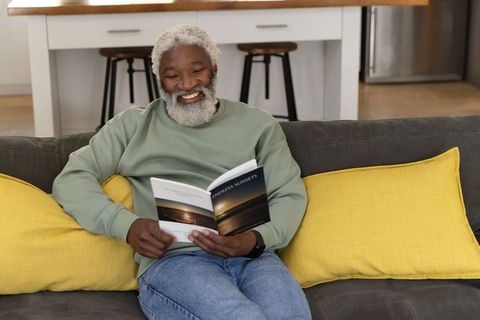 Senior man smiling while reading paperback on couch with bright yellow pillows