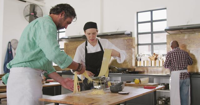 Professional chefs are making homemade pasta using fresh ingredients in a modern kitchen. One chef is cranking the pasta machine while another is assisting with the pasta strands. This image can be used for content related to cooking classes, culinary arts, teamwork in the kitchen, and promoting fresh homemade food.