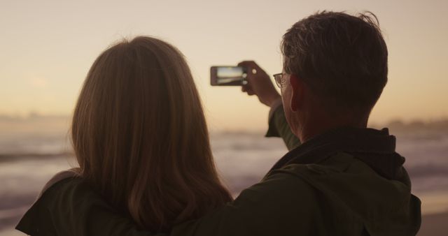 Senior caucasian couple taking pictures with smartphone on beach at sundown, copy space. Photography, relationship, retirement, romance, vacations, nature and active senior lifestyle, unaltered.