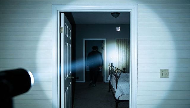 Silhouetted figure walking through dark hallway toward bedroom illuminated by flashlight at night