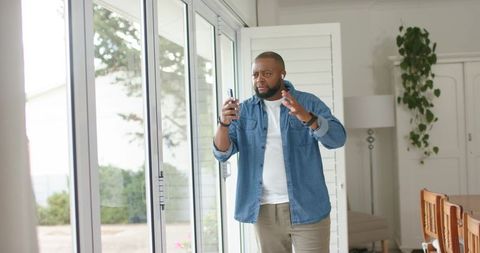 African man standing by sliding glass doors holding smartphone and gesturing on video call