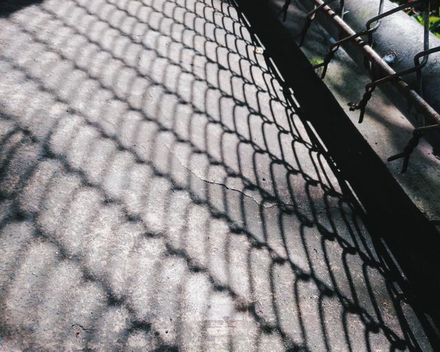 Shadow of a chain link fence creating a geometric pattern on a concrete surface. Sunlight casting intricate details, showcasing industrial and urban themes. Ideal for backgrounds, textures, urban projects, or design concepts involving patterns and shadows.